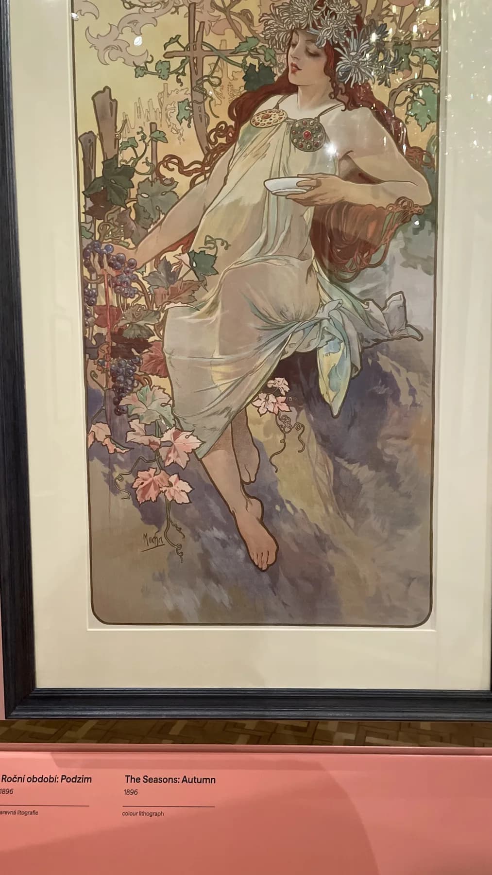 Alphonse Mucha's The Seasons: Autumn displayed in a museum frame, with the reclining figure, foliage, and lower label visible.